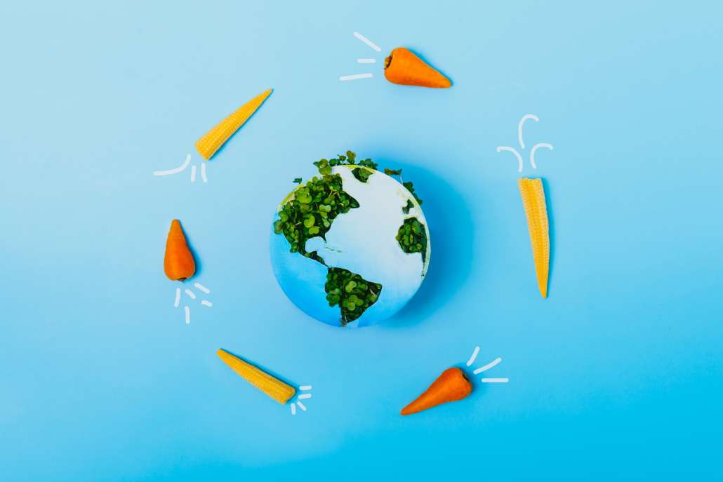 Creative veganism concept with rockets of carrots and baby corn flying around planet Earth, Earth model made of paper and fresh green sprouts collage on blue background.
