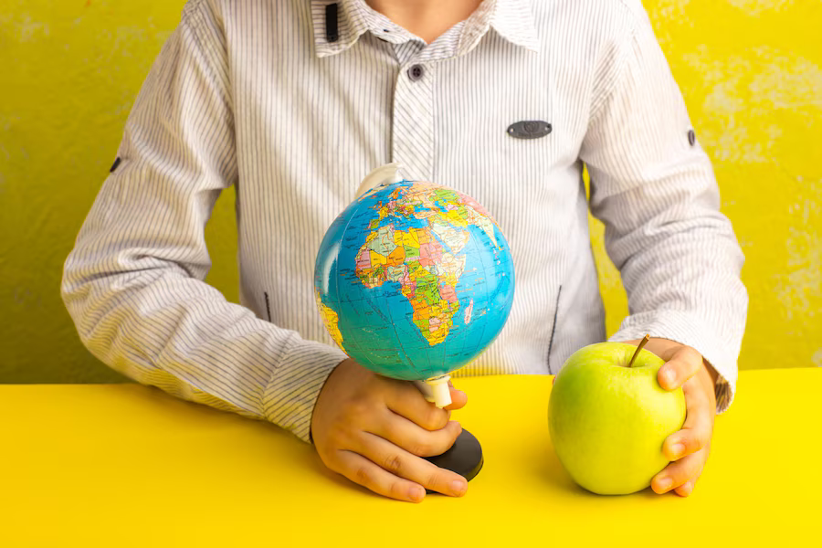 Front view of a little kid holding a little globe and a green apple on a yellow surface.