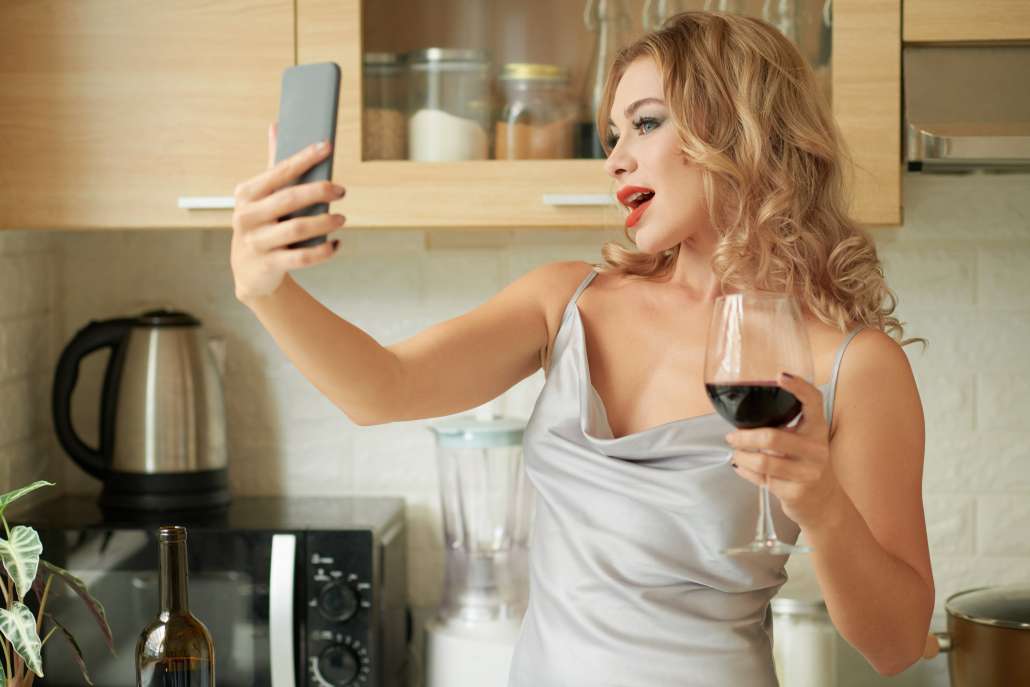 Young female influencer taking selfie with glass of wine in her kitchen